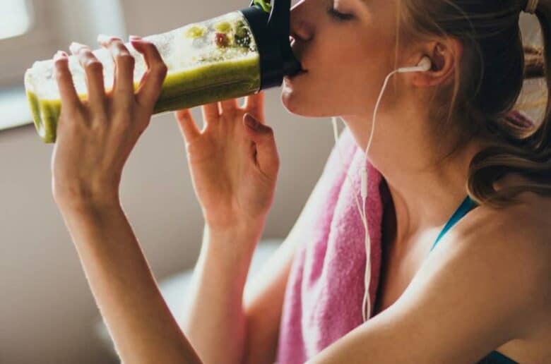 A woman wearing fitness apparel drinking a green smoothie. She has a pink towel over her shoulder and is wearing headphones.