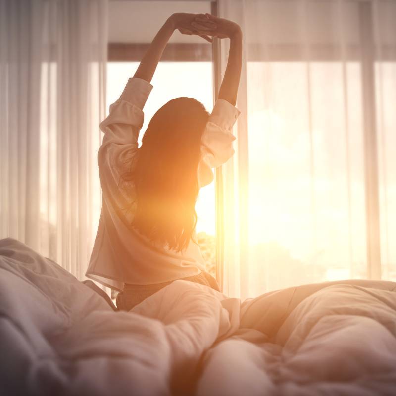 A woman just waking up to the sunrise coming through her window. She is stretching her arms over her head.