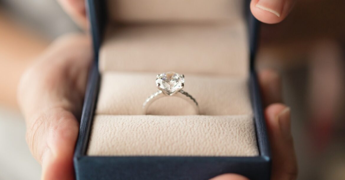 Hands hold an open navy ring box displaying a solitaire diamond engagement ring set against soft beige fabric.