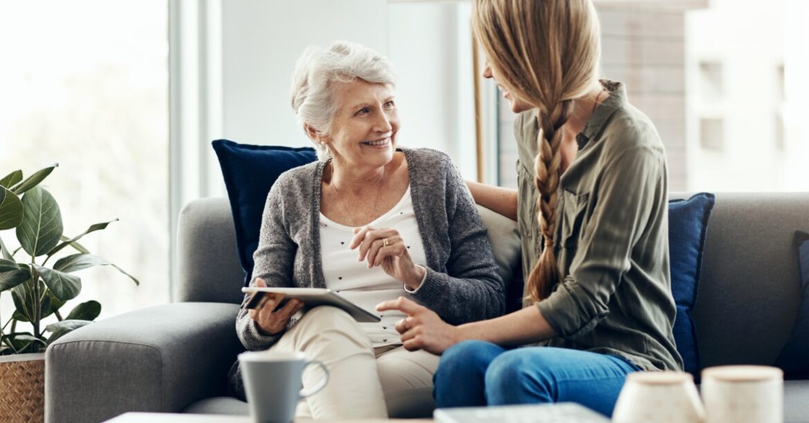 A smiling older woman sits on a couch with a tablet on her lap. A younger woman sits next to her with a hand on her shoulder.